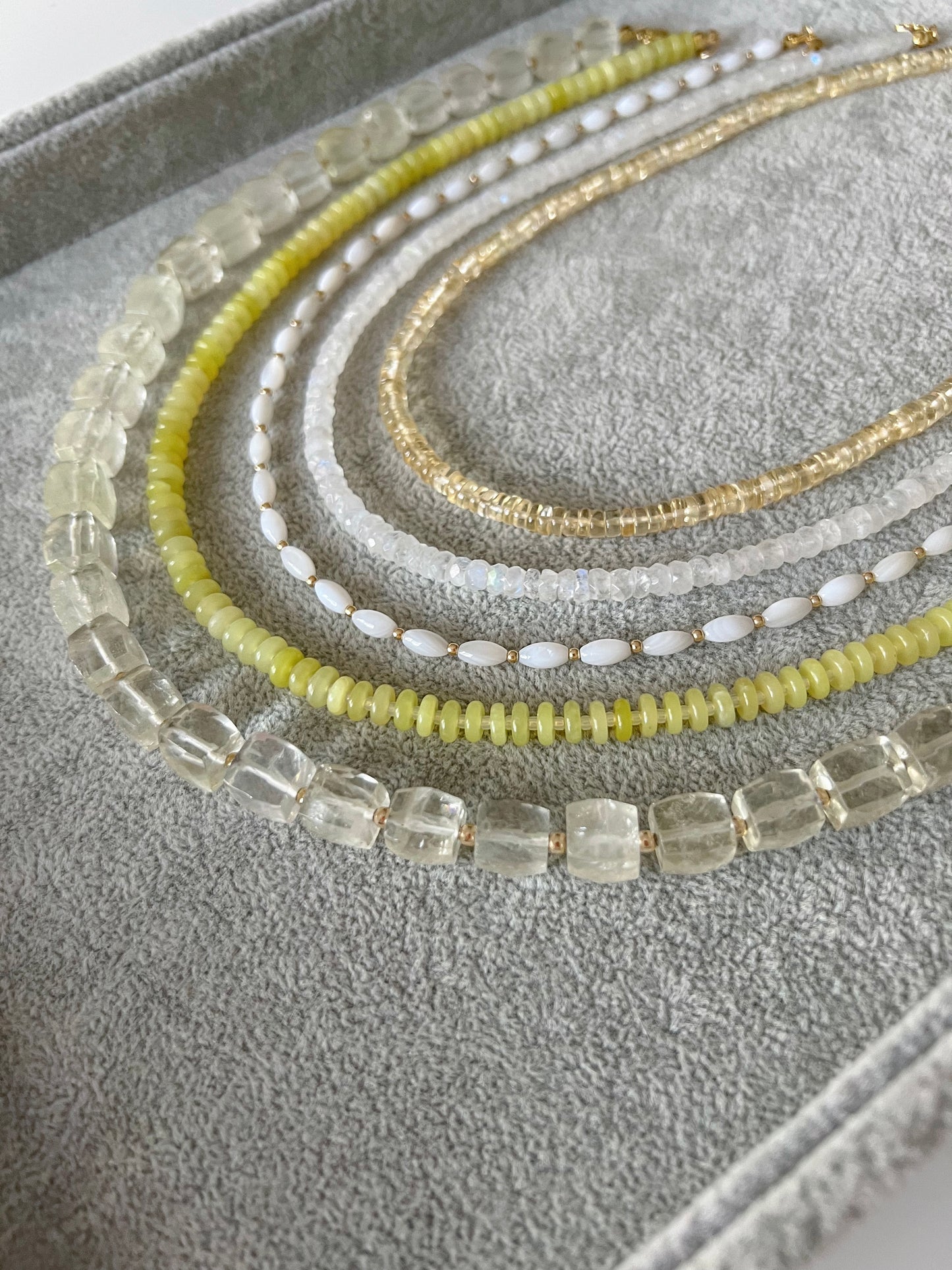 Five gemstone beaded necklaces on a grey velvet tray: citrine smooth rondelle, rainbow moonstone faceted rondelle, white shell rice shaped beads with gold accents, lemon jade smooth disc rondelle, and lemon quartz faceted cube with 14k solid gold or gold filled clasps