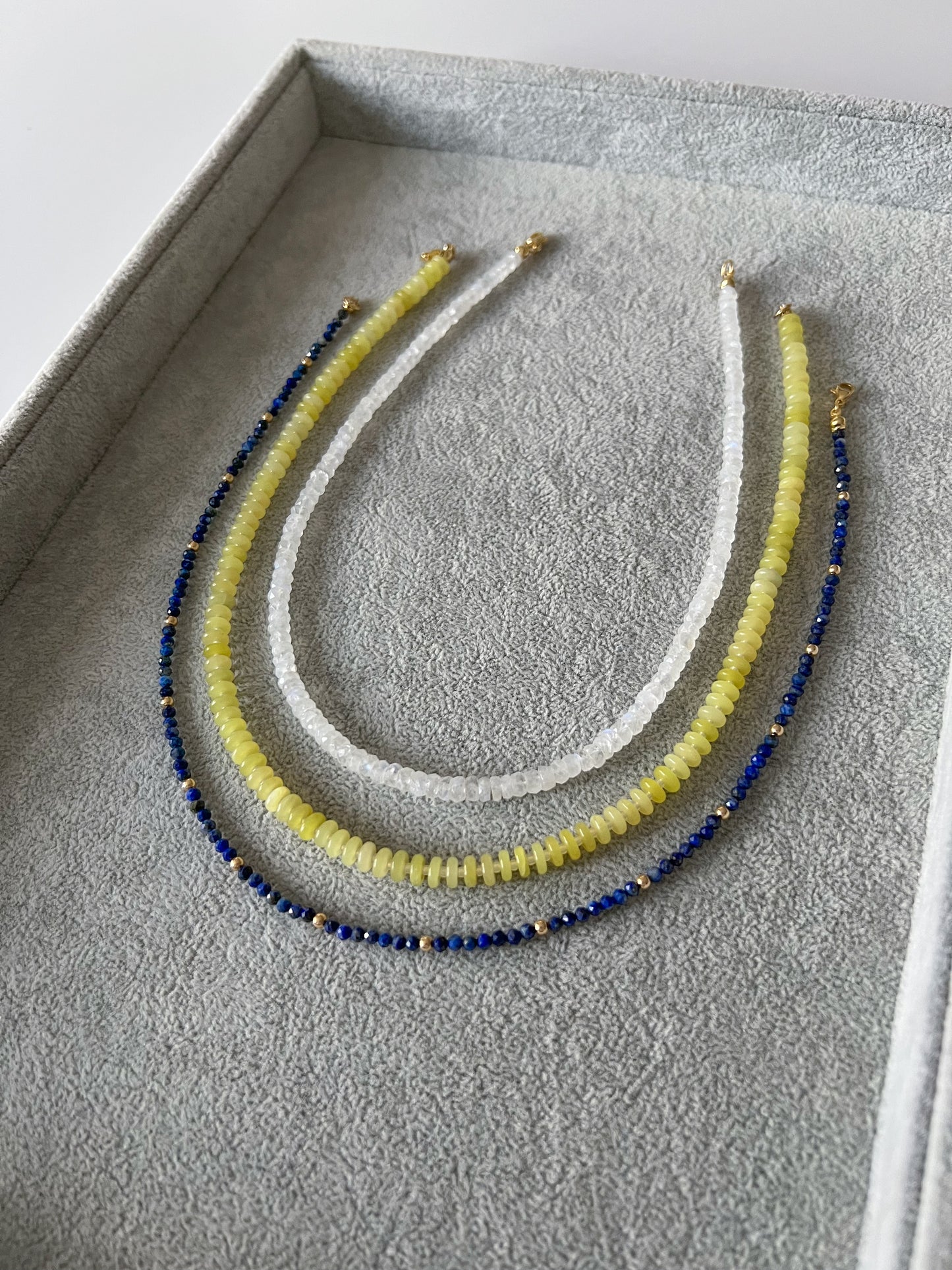 Three gemstone beaded necklaces on a grey velvet tray: rainbow moonstone faceted rondelle, lemon jade smooth disc rondelle, and lapis lazuli faceted round with 14k solid gold or gold filled accents and gold filled clasps