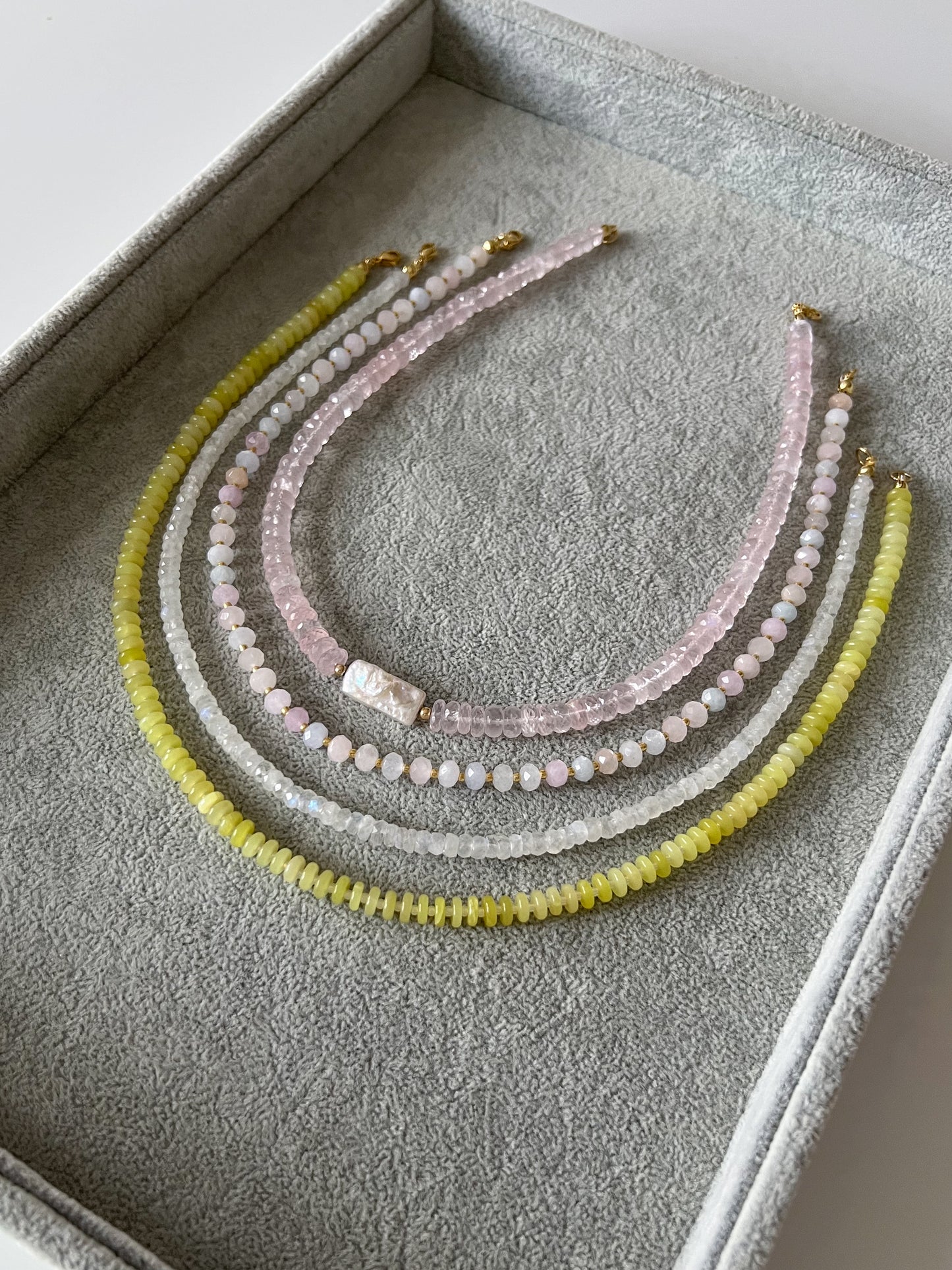 Four gemstone beaded necklaces on a gray velvet tray: rose quartz faceted rondelle with keshi pearl center, pastel morganite faceted rondelle, rainbow moonstone faceted rondelle, and lemon jade smooth disc rondelle with solid gold or 14k gold filled clasps