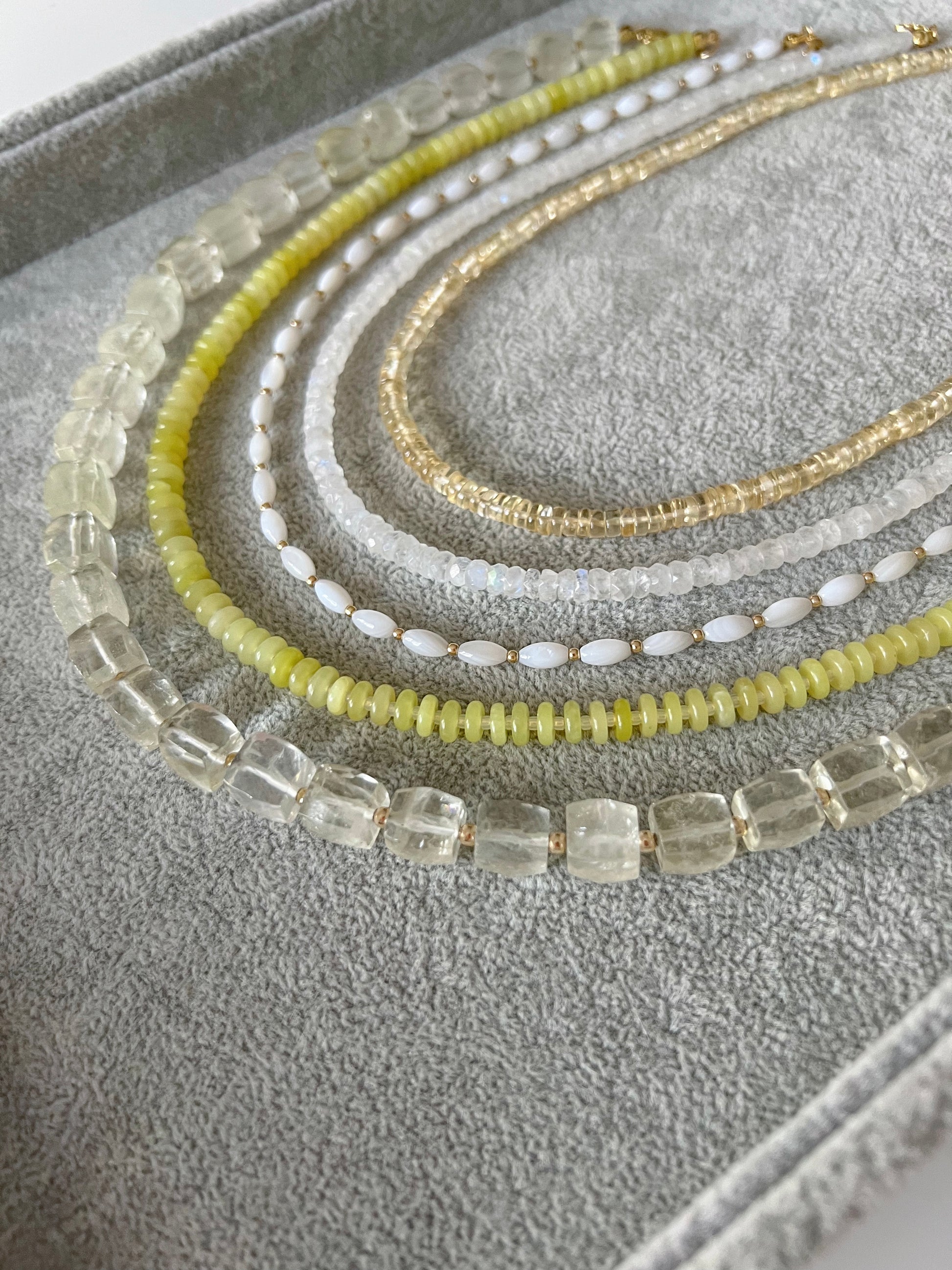 Five gemstone beaded necklaces on a grey velvet tray: citrine smooth rondelle, rainbow moonstone faceted rondelle, white shell rice shaped beads with gold accents, lemon jade smooth disc rondelle, and lemon quartz faceted cube with 14k solid gold or gold filled clasps
