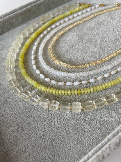 Five gemstone beaded necklaces on a grey velvet tray: citrine smooth rondelle, rainbow moonstone faceted rondelle, white shell rice shaped beads with gold accents, lemon jade smooth disc rondelle, and lemon quartz faceted cube with 14k solid gold or gold filled clasps