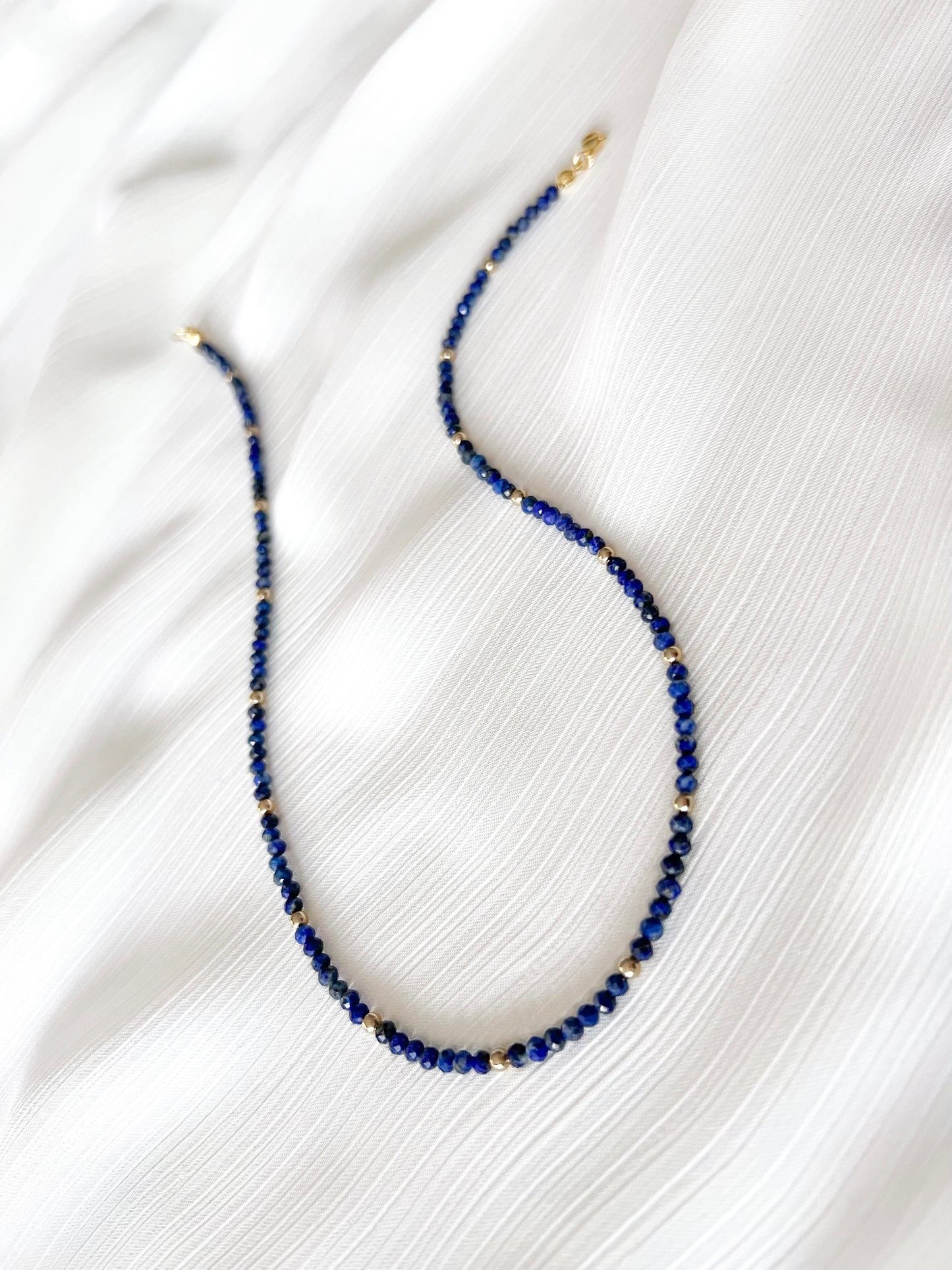 Dainty lapis lazuli beaded gemstone necklace with blue faceted round beads finished with gold accent beads and clasp on a soft white fabric background