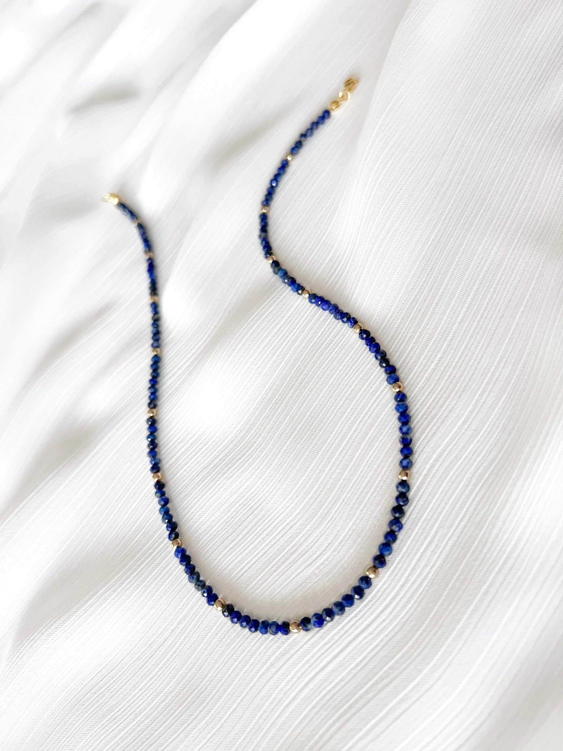 Dainty lapis lazuli beaded gemstone necklace with blue faceted round beads finished with gold accent beads and clasp on a soft white fabric background