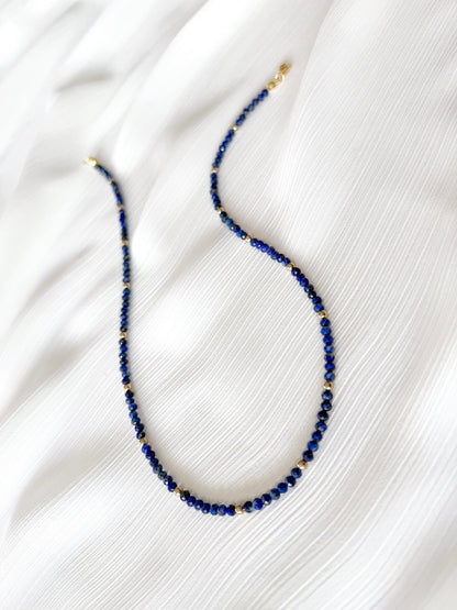 Dainty lapis lazuli beaded gemstone necklace with blue faceted round beads finished with gold accent beads and clasp on a soft white fabric background