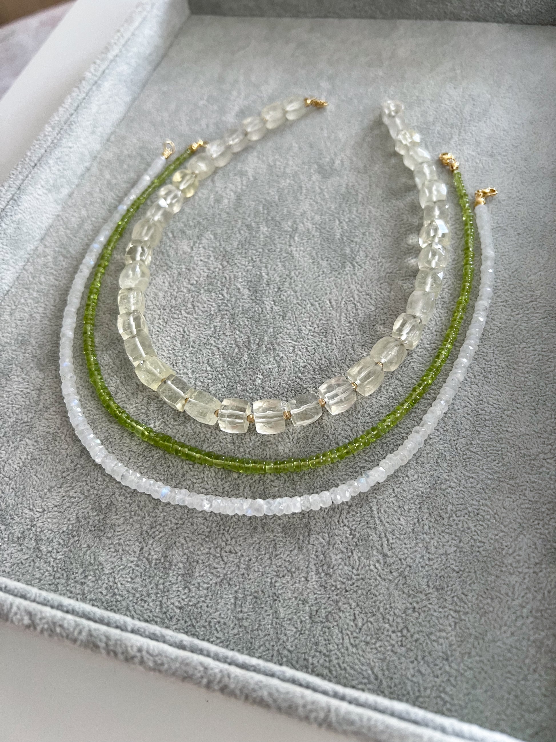 Three gemstone necklaces on a grey velvet tray: chunky lemon quartz cubes with gold filled accent beads, peridot smooth rondelle, and moonstone faceted rondelle, all with 14k solid gold or gold filled clasps