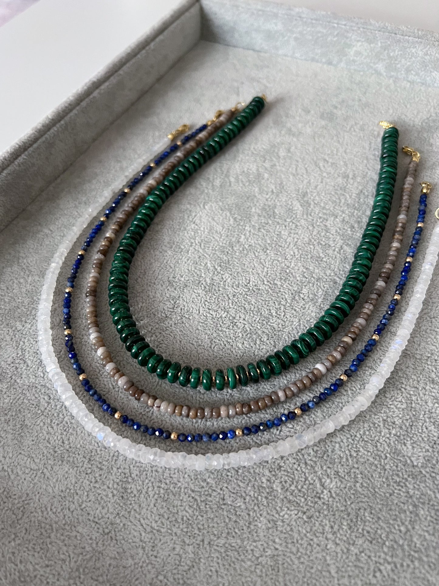 Four gemstone beaded necklaces on a grey velvet tray: chunky malachite disc rondelles with gold accent beads, silver leaf jasper smooth rondelle, lapis lazuli  with gold accents, and rainbow moonstone faceted rondelle with 14k solid gold or gold filled clasps