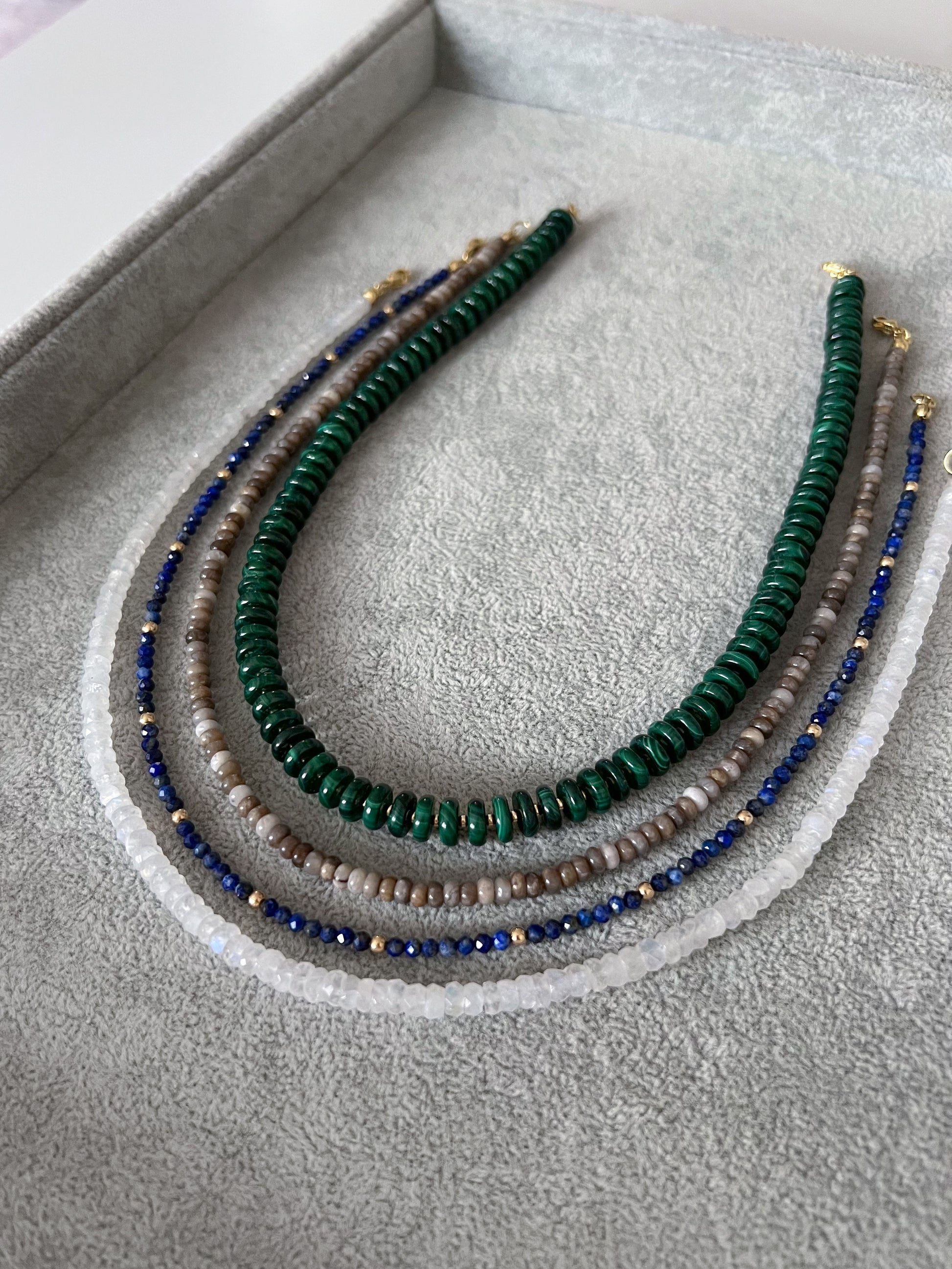 Four gemstone beaded necklaces on a grey velvet tray: chunky malachite disc rondelles with gold accent beads, silver leaf jasper smooth rondelle, lapis lazuli  with gold accents, and rainbow moonstone faceted rondelle with 14k solid gold or gold filled clasps