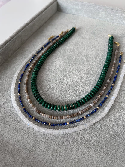 Four gemstone beaded necklaces on a grey velvet tray: chunky malachite disc rondelles with gold accent beads, silver leaf jasper smooth rondelle, lapis lazuli  with gold accents, and rainbow moonstone faceted rondelle with 14k solid gold or gold filled clasps