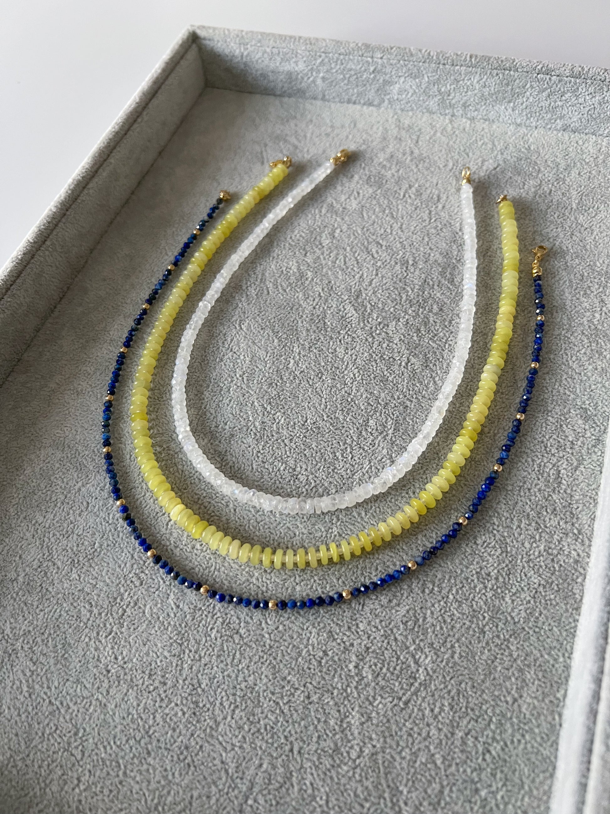 Three gemstone beaded necklaces on a grey velvet tray: rainbow moonstone faceted rondelle, lemon jade smooth disc rondelle, and lapis lazuli faceted round with 14k solid gold or gold filled accents and gold filled clasps