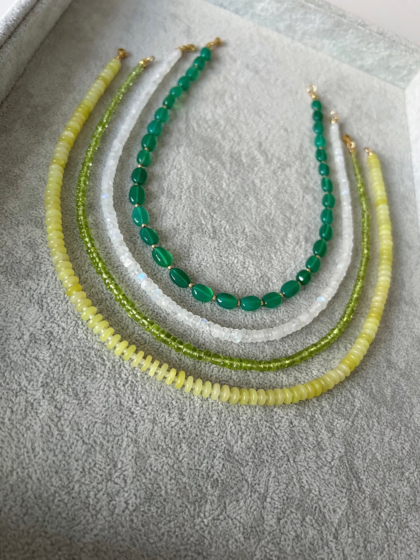 Four gemstone beaded necklaces on a grey velvet tray: green onyx smooth ovals with gold accent beads, rainbow moonstone faceted rondelle, peridot smooth rondelle, and lemon jade smooth disc rondelle with 14k gold filled clasps