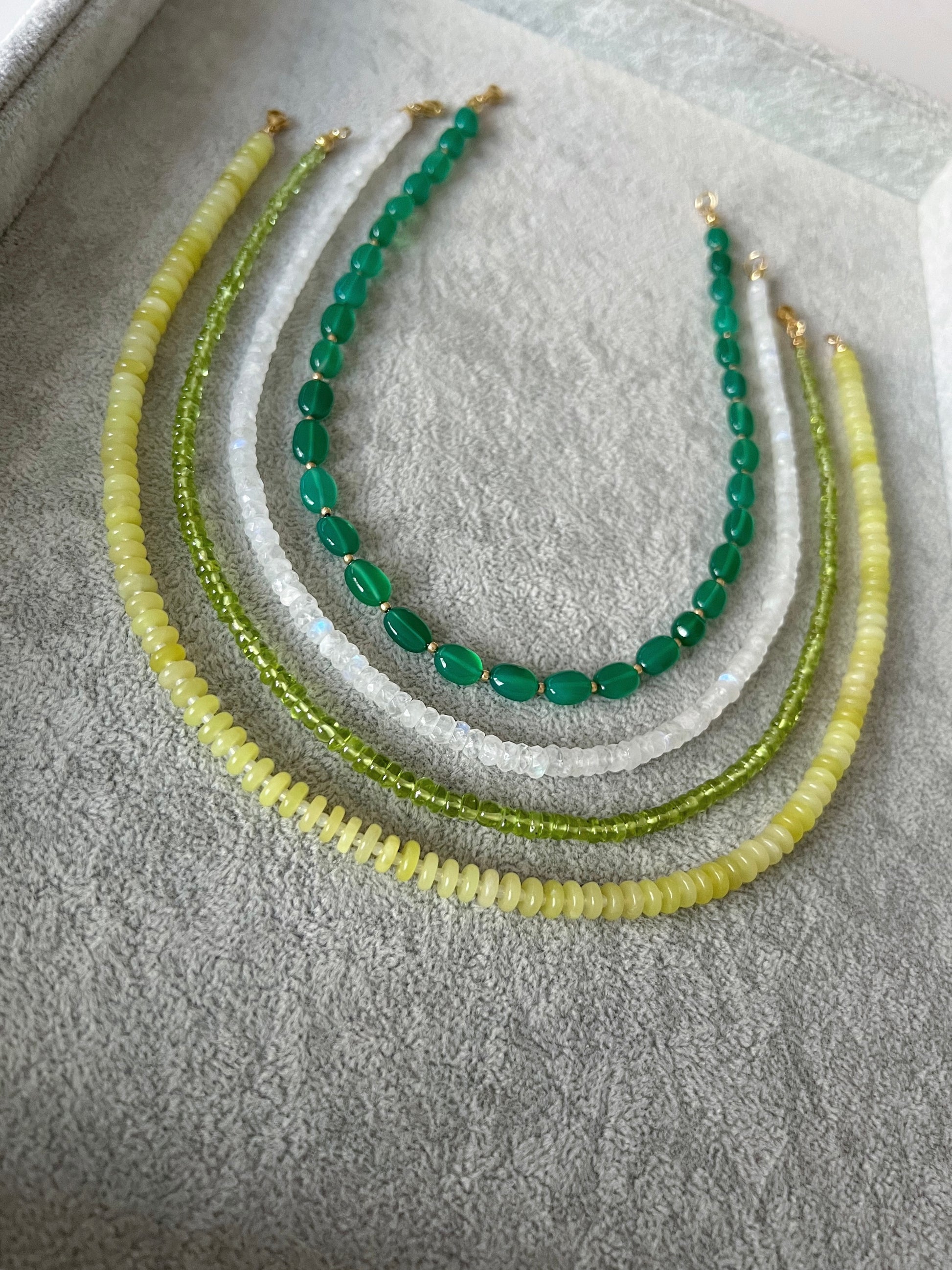 Four gemstone beaded necklaces on a grey velvet tray: green onyx smooth ovals with gold accent beads, rainbow moonstone faceted rondelle, peridot smooth rondelle, and lemon jade smooth disc rondelle with 14k gold filled clasps