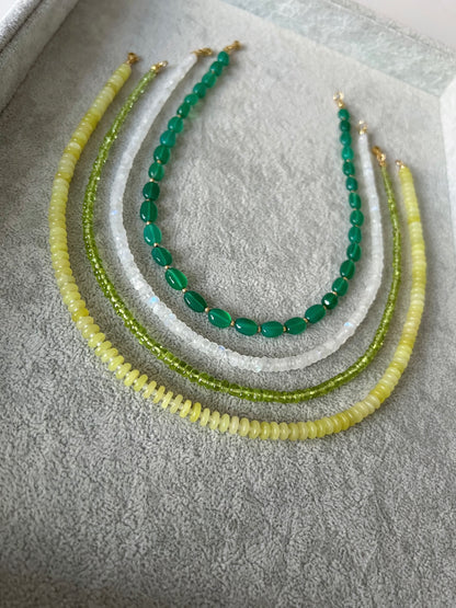 Four gemstone beaded necklaces on a grey velvet tray: green onyx smooth ovals with gold accent beads, rainbow moonstone faceted rondelle, peridot smooth rondelle, and lemon jade smooth disc rondelle with 14k gold filled clasps