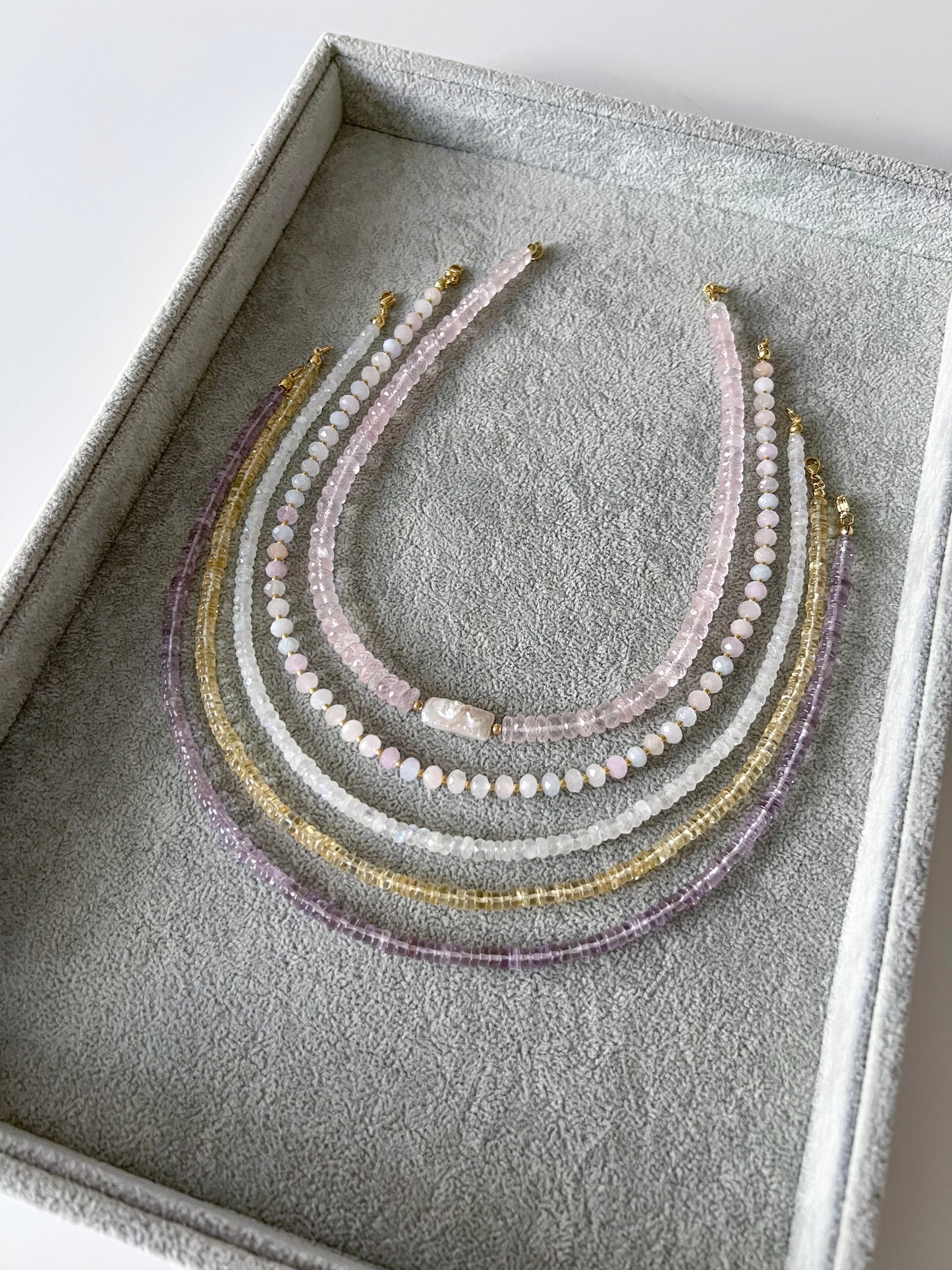 Four gemstone necklaces on a grey velvet tray: chunky faceted rose quartz with a freshwater keshi pearl centerpiece, morganite with faceted rondelle beads and gold filled accents, rainbow moonstone faceted rondelle, citrine smooth rondelle, and pink amethyst smooth rondelle