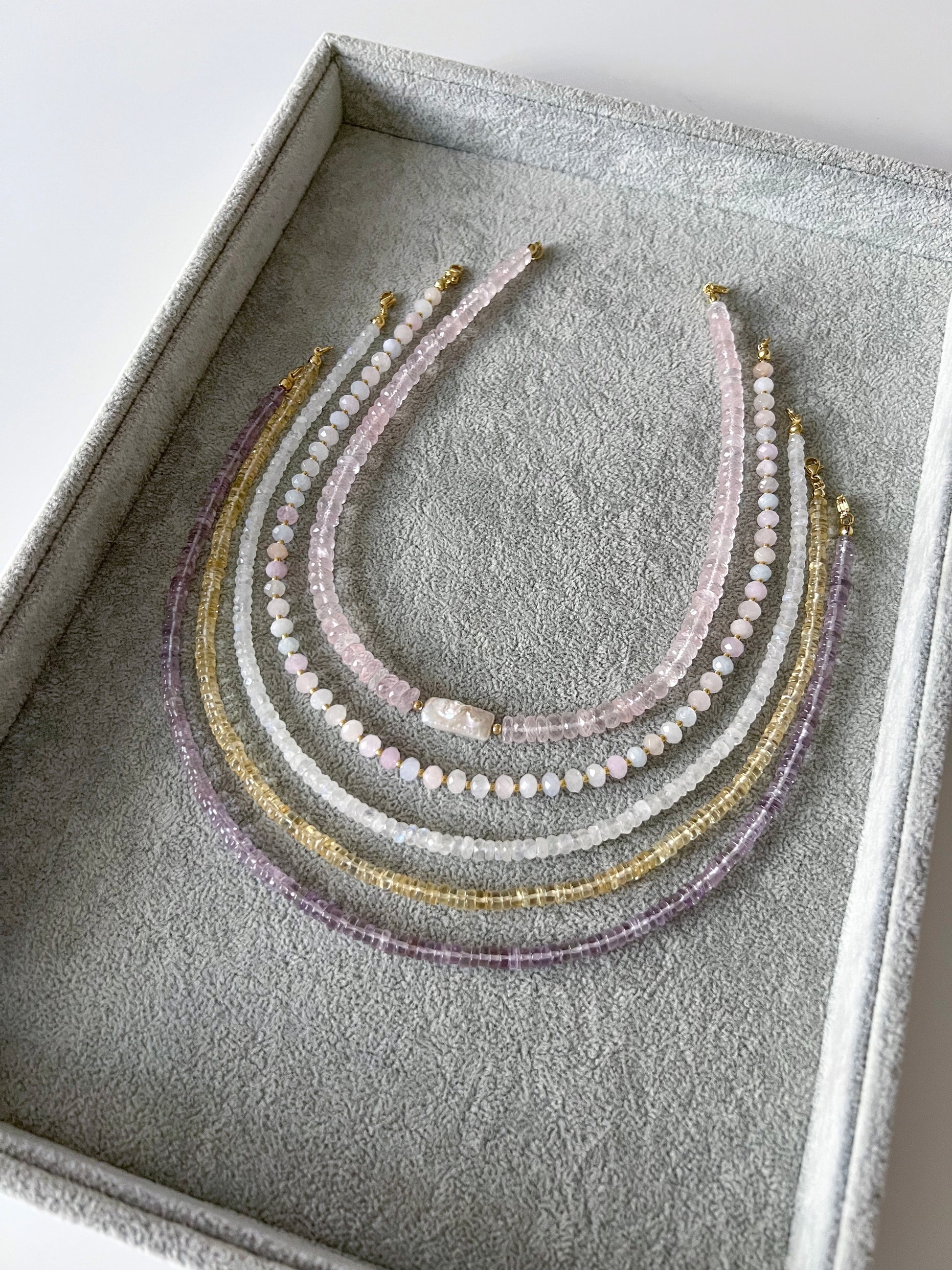 Four gemstone necklaces on a grey velvet tray: chunky faceted rose quartz with a freshwater keshi pearl centerpiece, morganite with faceted rondelle beads and gold filled accents, rainbow moonstone faceted rondelle, citrine smooth rondelle, and pink amethyst smooth rondelle