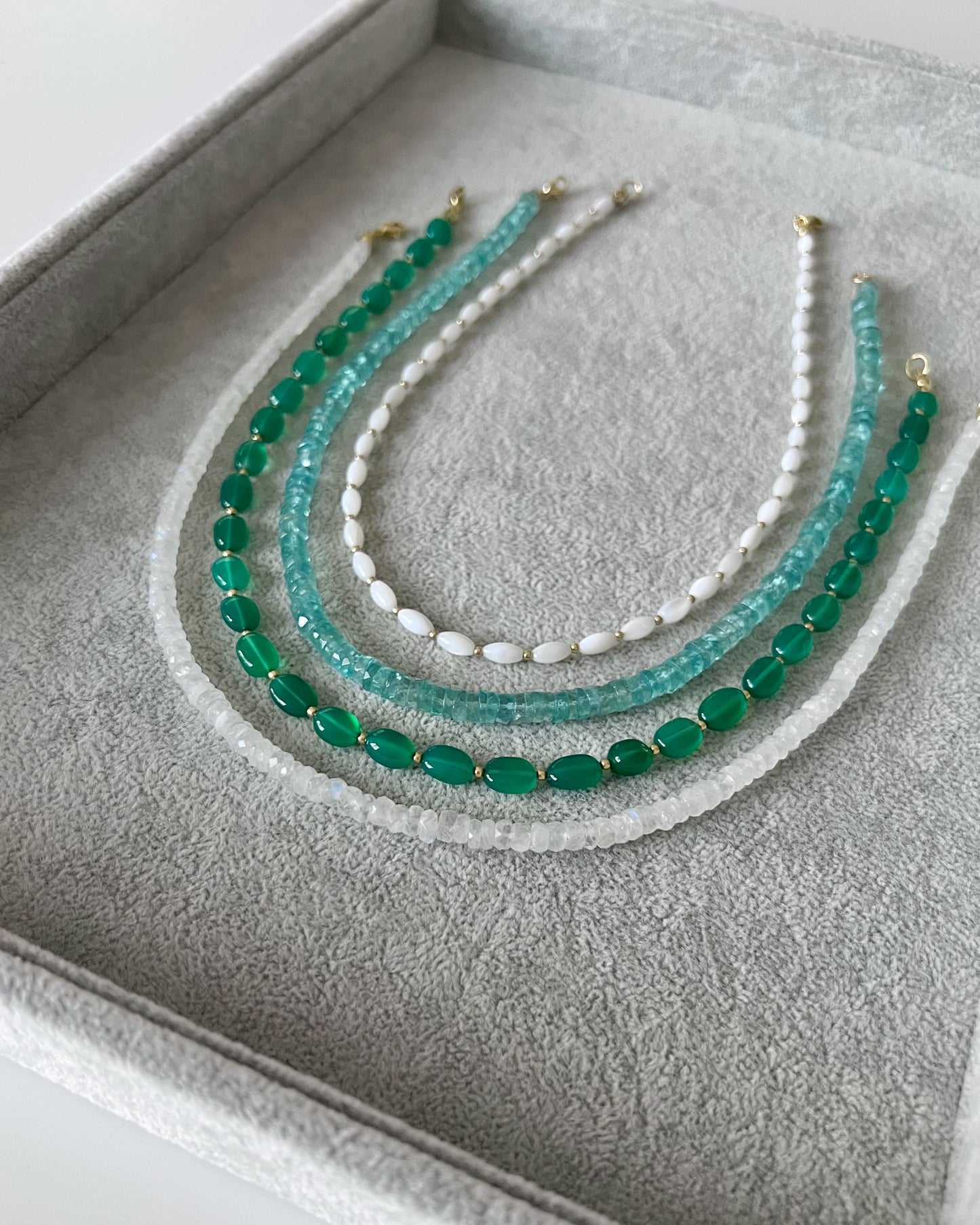 Four gemstone beaded necklaces on a grey velvet tray: white shell smooth rice shaped with gold accents, sky blue apatite faceted heishi, green onyx oval with gold accents, and rainbow moonstone faceted rondelle finished with 14k solid gold or gold filled clasps