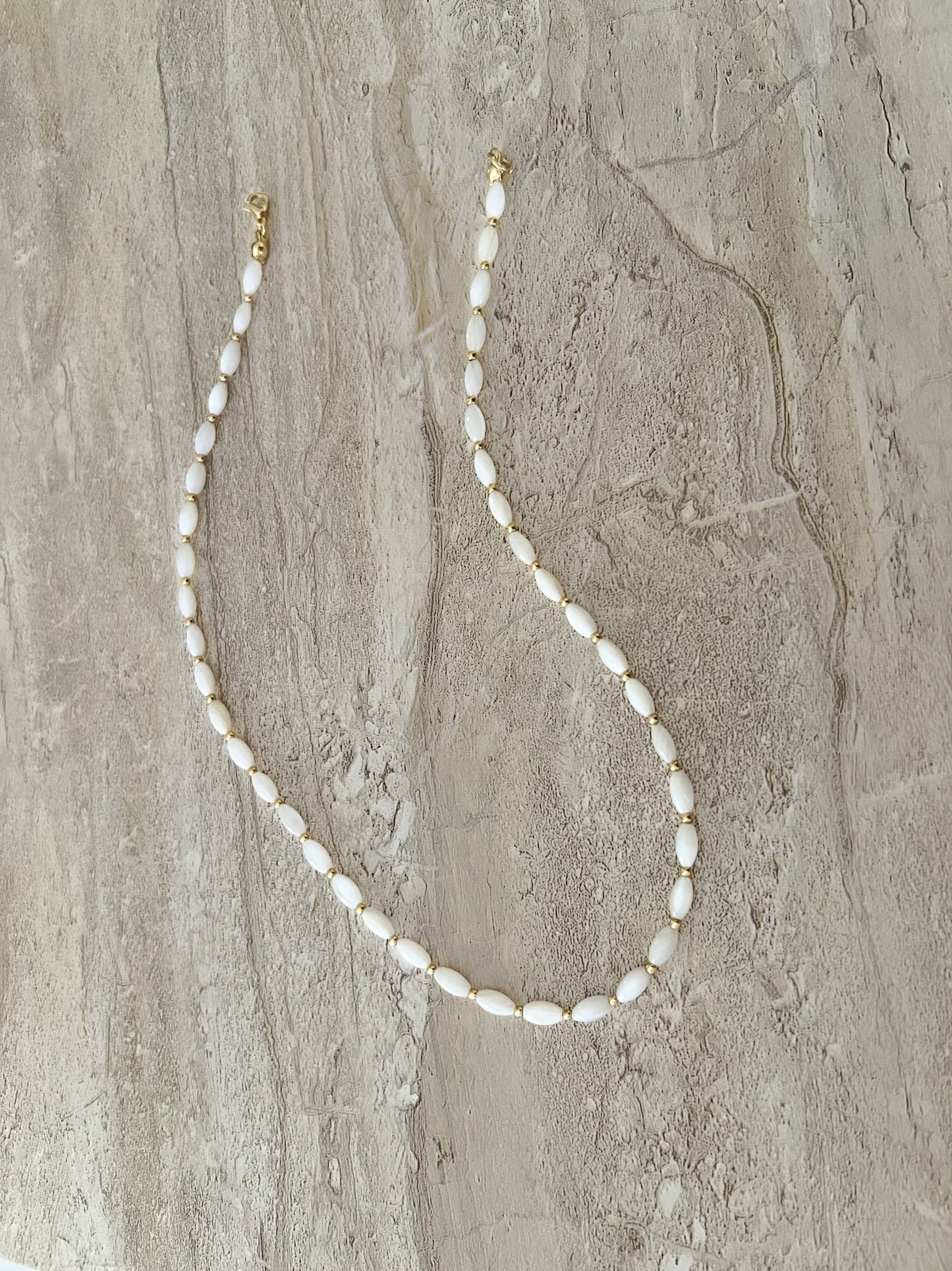 Dainty natural shell beaded necklace with white rice shaped shell beads and 14k gold filled accent beads and clasp. Minimalist layering necklace on a luxury stone slab background