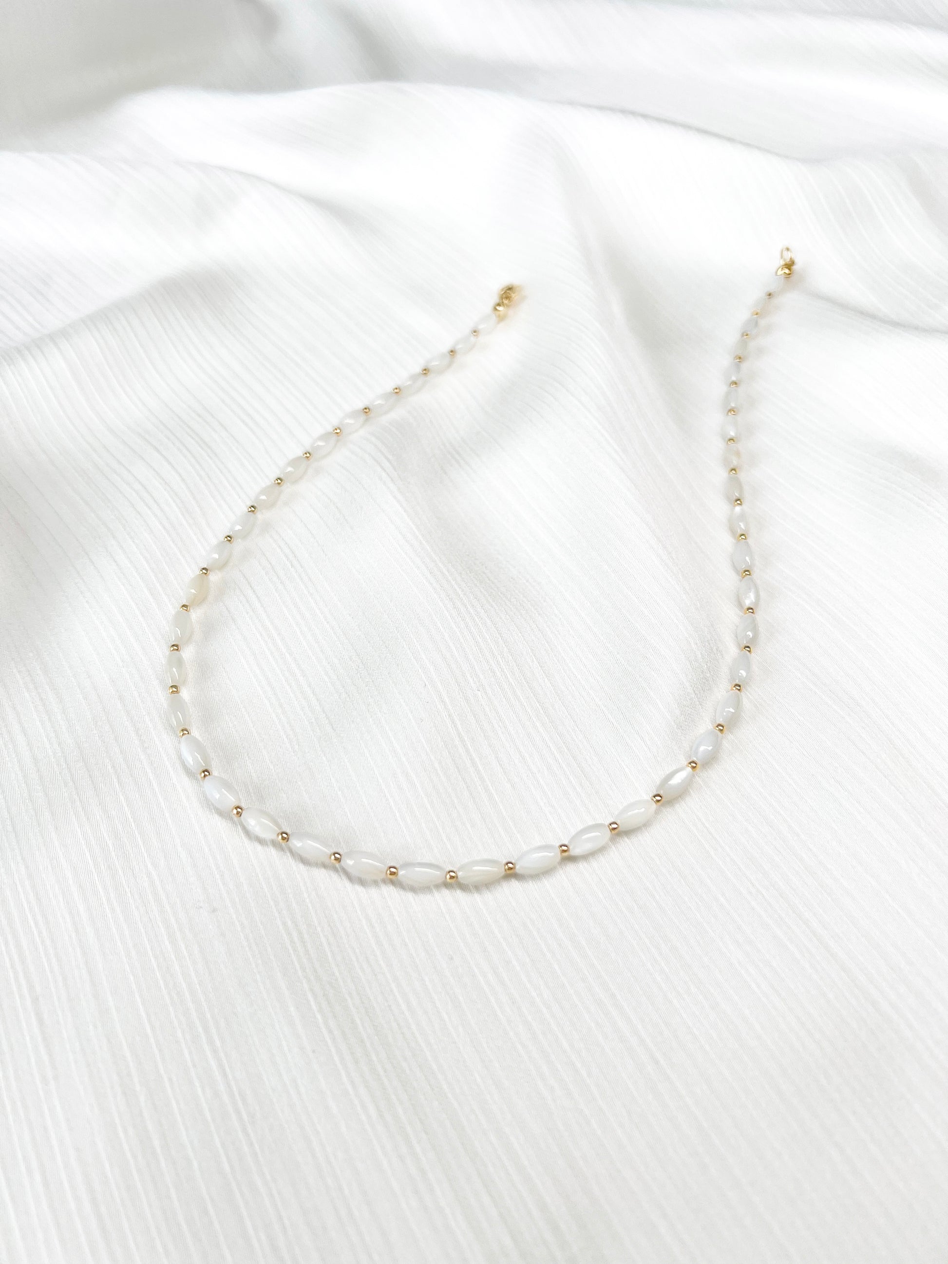 Dainty natural shell beaded necklace with white rice shaped shell beads and 14k gold filled accent beads and clasp. Minimalist layering necklace on a soft white fabric background