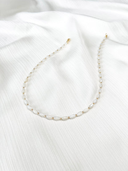 Dainty natural shell beaded necklace with white rice shaped shell beads and 14k gold filled accent beads and clasp. Minimalist layering necklace on a soft white fabric background